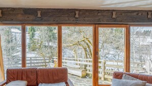 A cozy room with a brown leather sofa and a wooden bench, large windows with a view of a snowy outdoor area and a wooden fence.