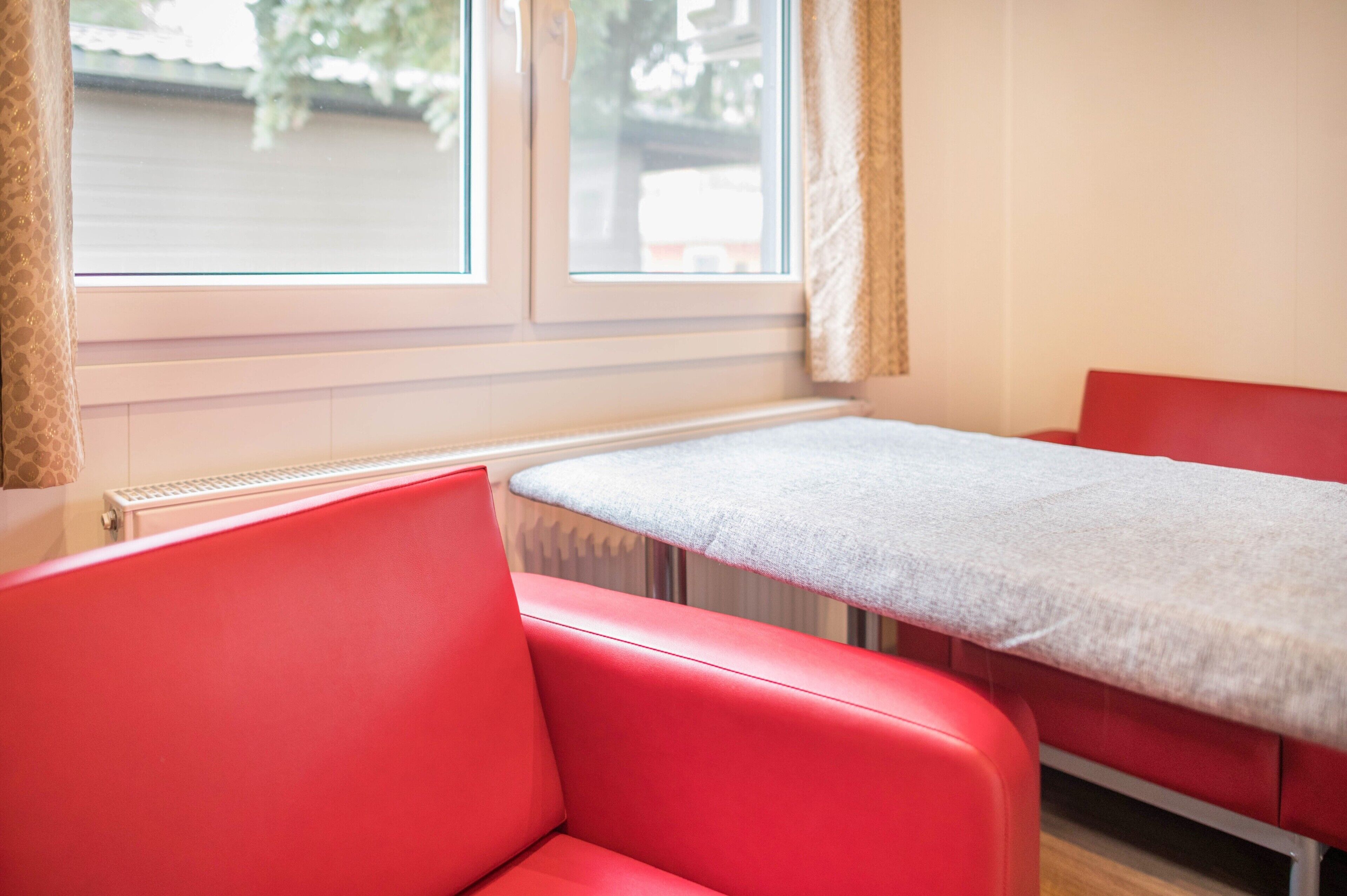 A room with a red chair, a table with a grey cloth, and a window with curtains.
