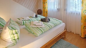 A room with two beds, wooden flooring, a hanging light fixture, and a window with curtains.