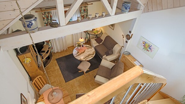 A loft-style living area with a high ceiling, wooden beams, a sofa set, a dining table, and a kitchen area with a stove.