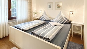 Una cama doble con ropa de cama a rayas, dos mesitas de noche, una mesita auxiliar, una ventana con cortinas y lámparas fijadas a la pared.
