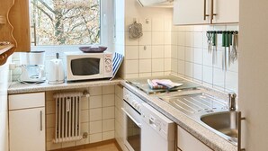 A kitchen with a microwave, kettle, and kettle on the counter. There is a window with a view of trees.