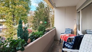A balcony with a view of trees and buildings, two chairs with checkered upholstery, and a table with a red cloth.
