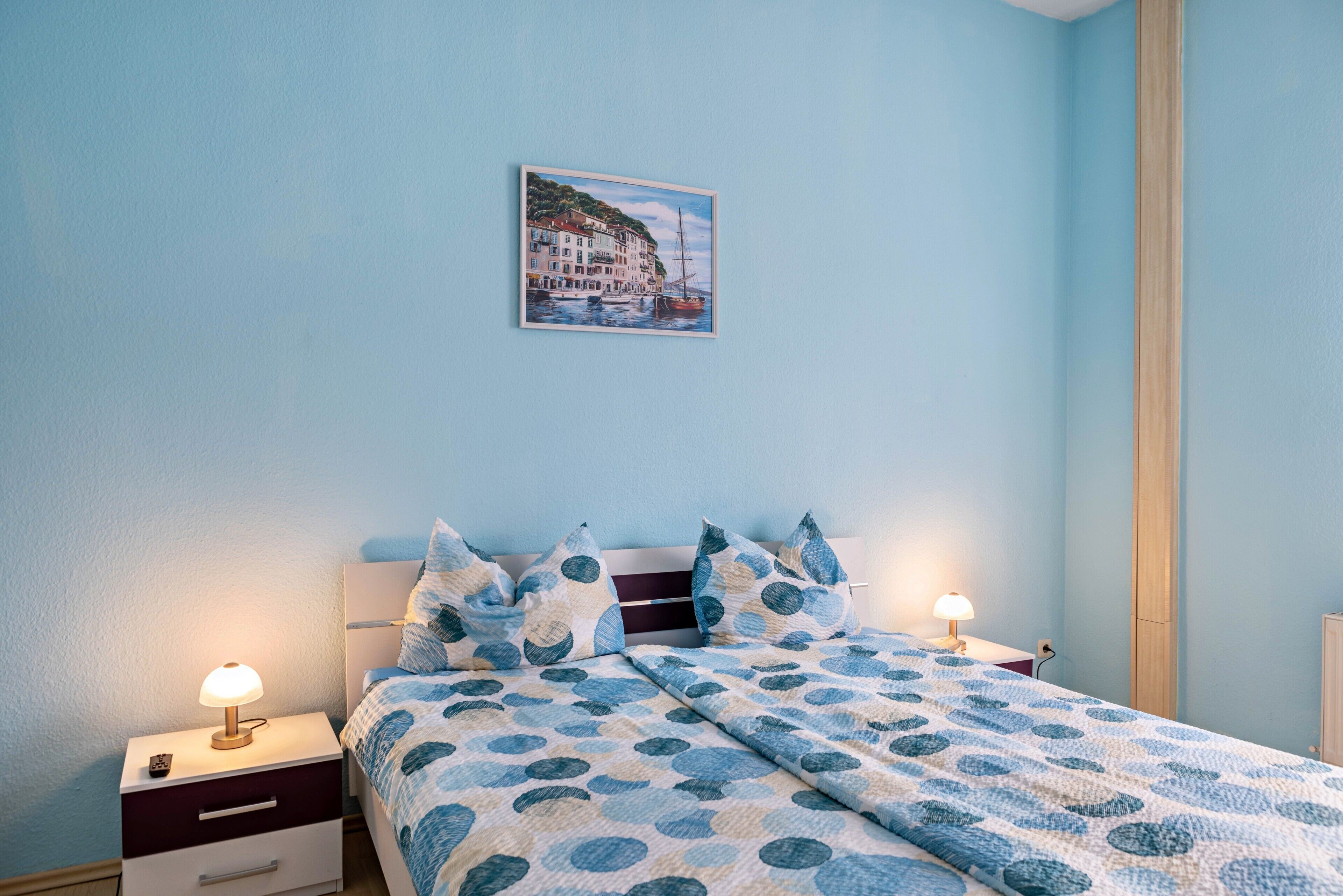 Ein Schlafzimmer mit einem Bett, zwei Nachttischen, einer Lampe, einem Wandbild und einem Fenster.