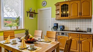 A kitchen with wooden cabinets, a window with lace curtains, a clock, and a dining table set with yellow napkins and fruit. - Apartment "Burgblick" with Mountain View, Shared Terrace & Wi-Fi (Plaue)