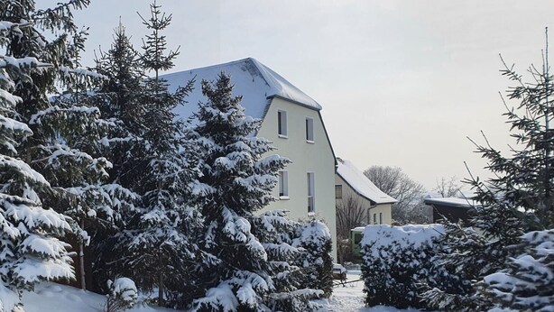 Paisatge cobert de neu amb una casa, arbres i un cel serè.