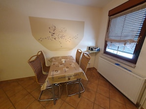 Un coin repas avec une table recouverte d’une nappe à motifs de café, quatre chaises en osier, une décoration murale représentant des grains de café et un cœur, une petite radio et une fenêtre avec des stores.