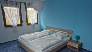 Un dormitori amb un llit de fusta, dues taules de nit, una tauleta auxiliar i una finestra amb cortines transparents.