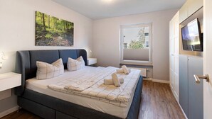 A bedroom with a bed, a nightstand, a wall-mounted TV, a window with a view, and a ceiling light fixture. - Apartment 'Hexe 4 ' with Balcony and Wi-Fi (Braunlage)