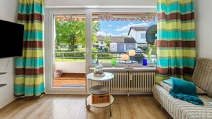 Een kamer met een bank, een klein tafeltje, een rieten mand, een radiator en uitzicht op een woonwijk.