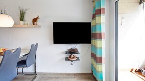 Een moderne woonkamer met een flatscreen televisie aan de muur, een eethoek met stoelen en een tafel, en een raam met een gestreept gordijn.