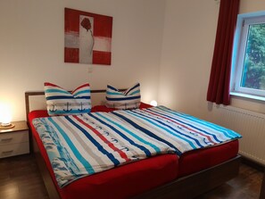 Un lit avec une literie à rayures, une table de chevet avec une lampe, un mur orné d’un tableau et une fenêtre avec des rideaux.