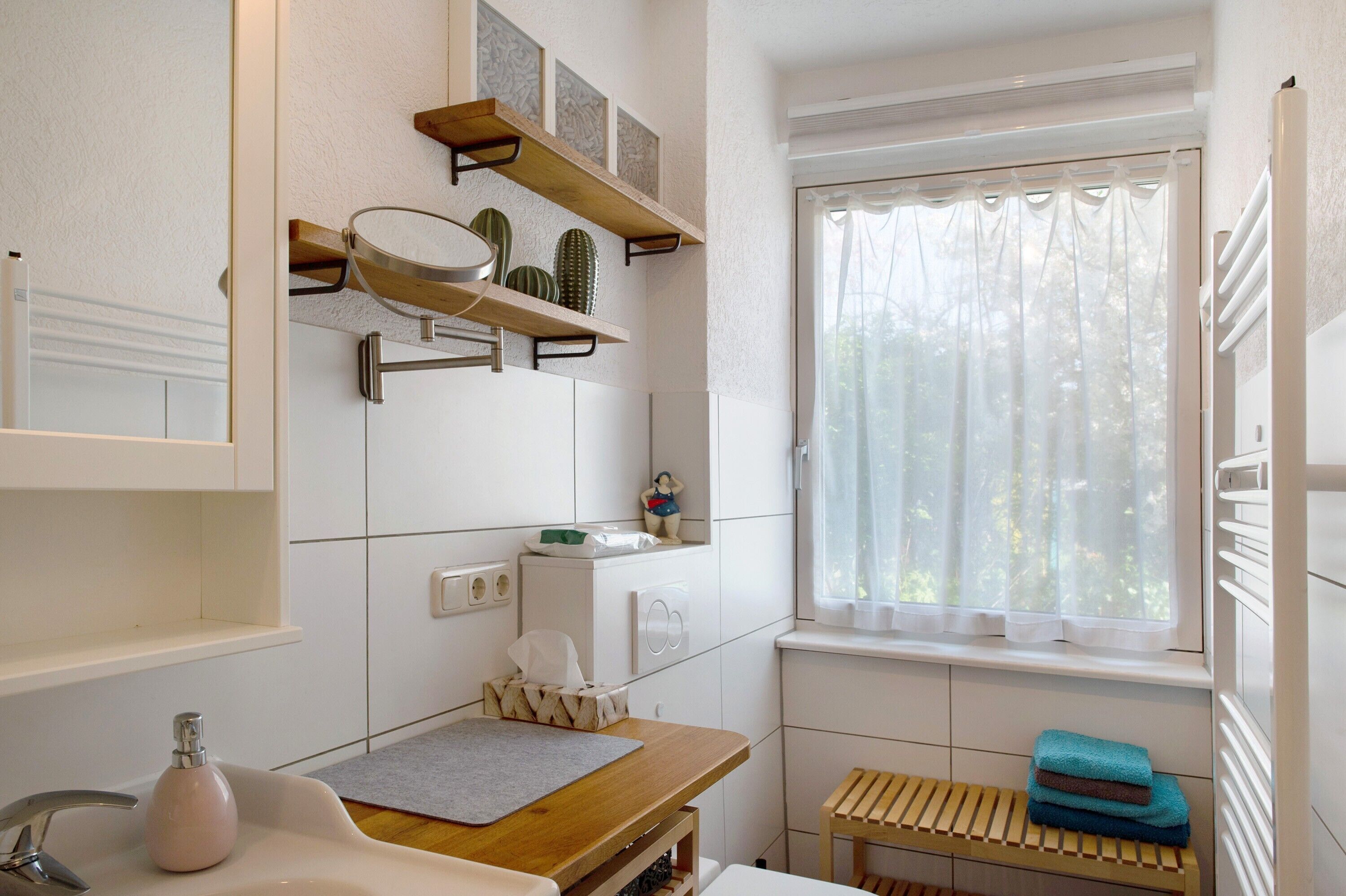 Een badkamer met een wastafel, spiegel, planken met decoratieve spullen, een raam met gordijnen en een bankje.