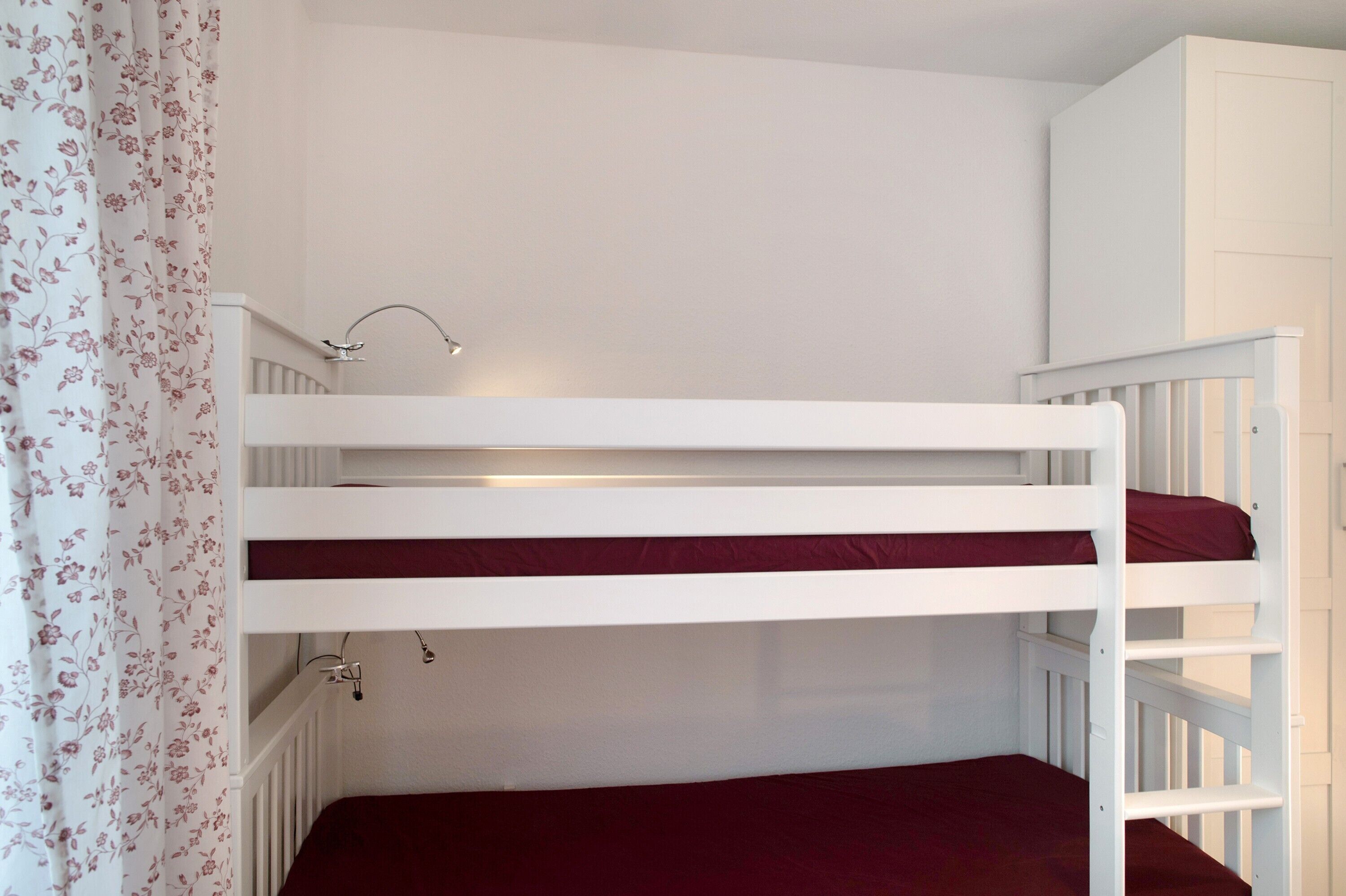 Una cama litera con colchón rojo, barandillas blancas y una lámpara pequeña en la pared.