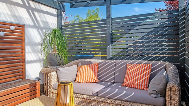 A wicker sofa with cushions, a small round table, and a wooden bench in a sunlit outdoor area with a pergola.