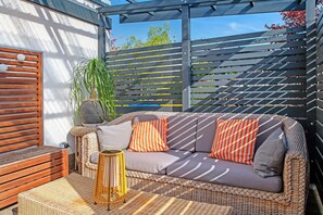 A wicker sofa with cushions, a small round table, and a wooden bench in a sunlit outdoor area with a pergola. - Holiday Apartment 'Alte Schule' with Private Terrace and Wi-Fi (Nienhagen)