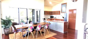 A dining area with a wooden table and chairs, a kitchen with wooden cabinets, and a large window. - BankSea Holiday Home (Agnes Water)