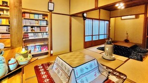 Ein traditioneller japanischer Raum mit Tatami-Böden, einem niedrigen Tisch mit Tischläufer, einem Bücherregal mit Büchern und DVDs und einem Shoji-Bildschirm mit Blick hinaus.
