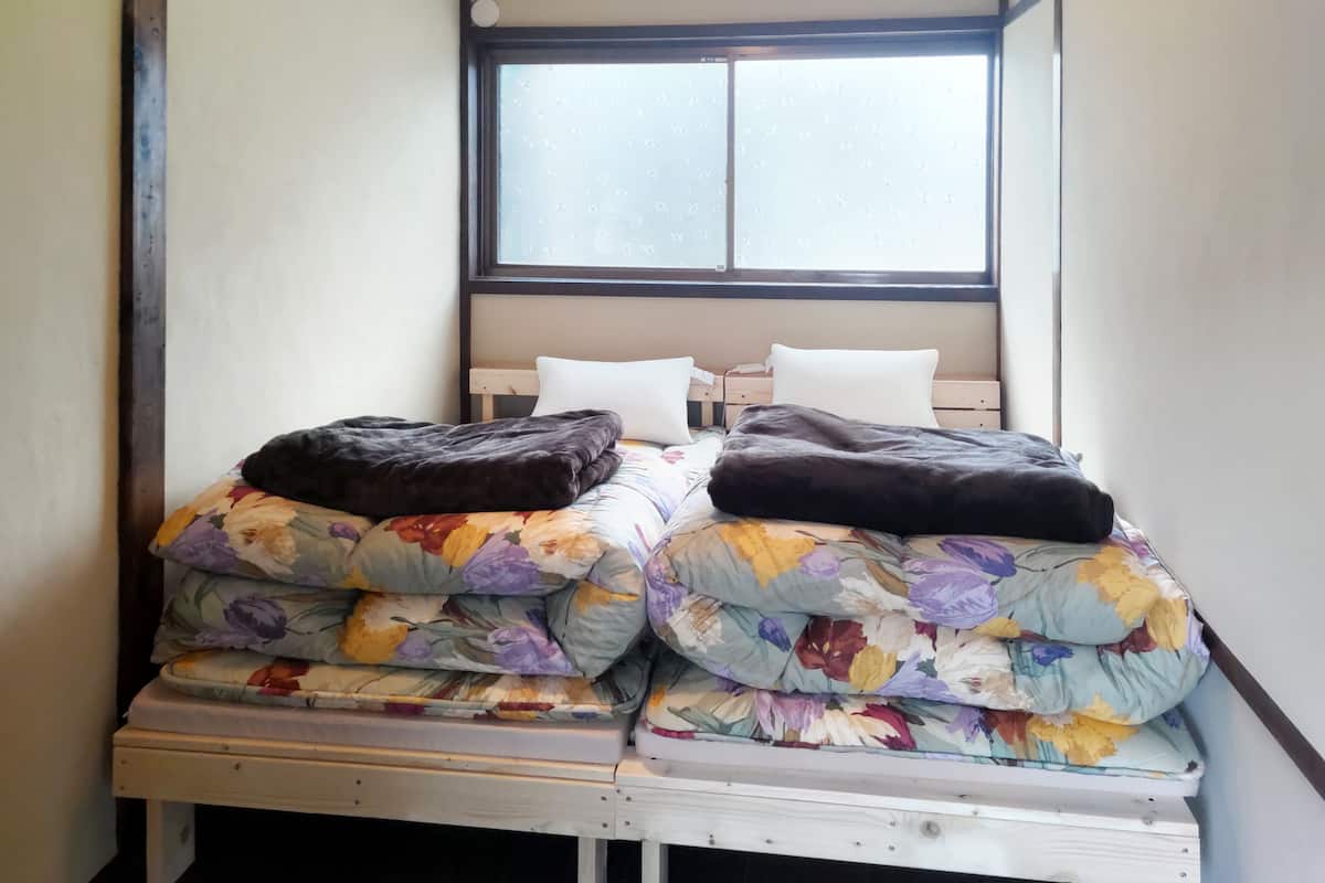 Una habitación pequeña con dos camas, cada una con ropa de cama floral y una manta oscura. Hay una ventana con vidrio esmerilado y una unidad de aire acondicionado blanca encima.