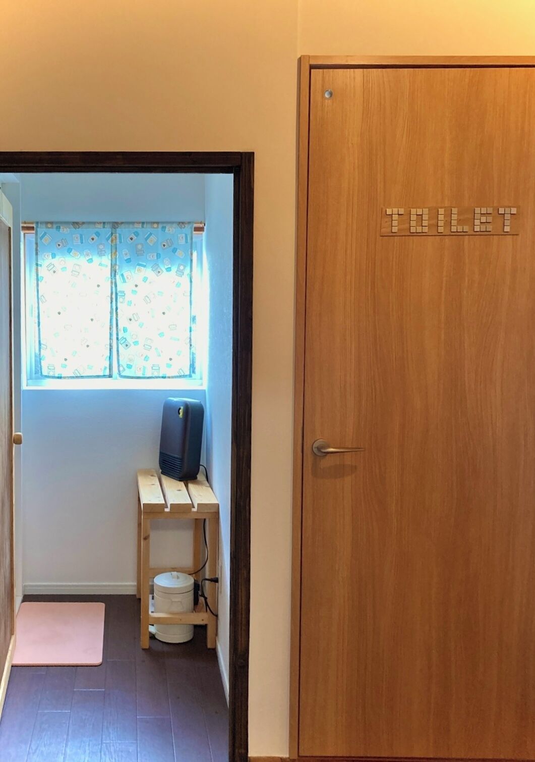 A wooden door with a toilet sign, a window with patterned curtains, a small desk with a computer, and a pink mat on the floor.