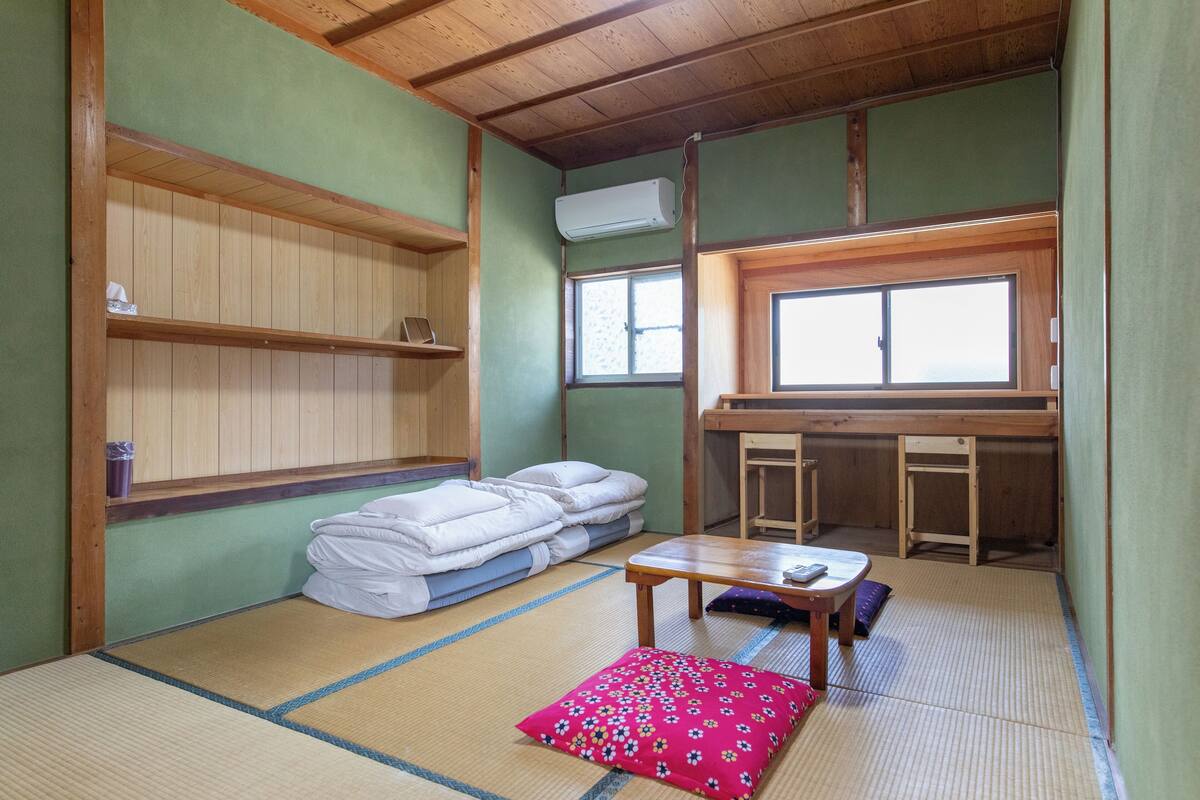 Una habitación japonesa tradicional con suelo de tatami, futón, una pequeña mesa de madera y ventana con vistas.
