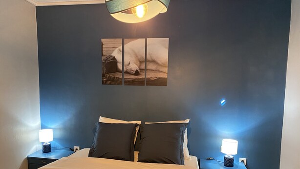 Un dormitorio con cama, dos mesitas de noche, una lámpara de pared e imagen enmarcada.