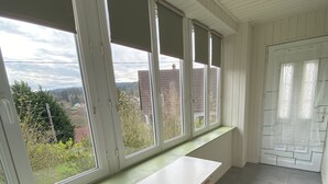 Una habitación con una larga mesa blanca y dos sillas de madera, ventanas amplias que dan a edificios y árboles, y una puerta con diseño decorativo.