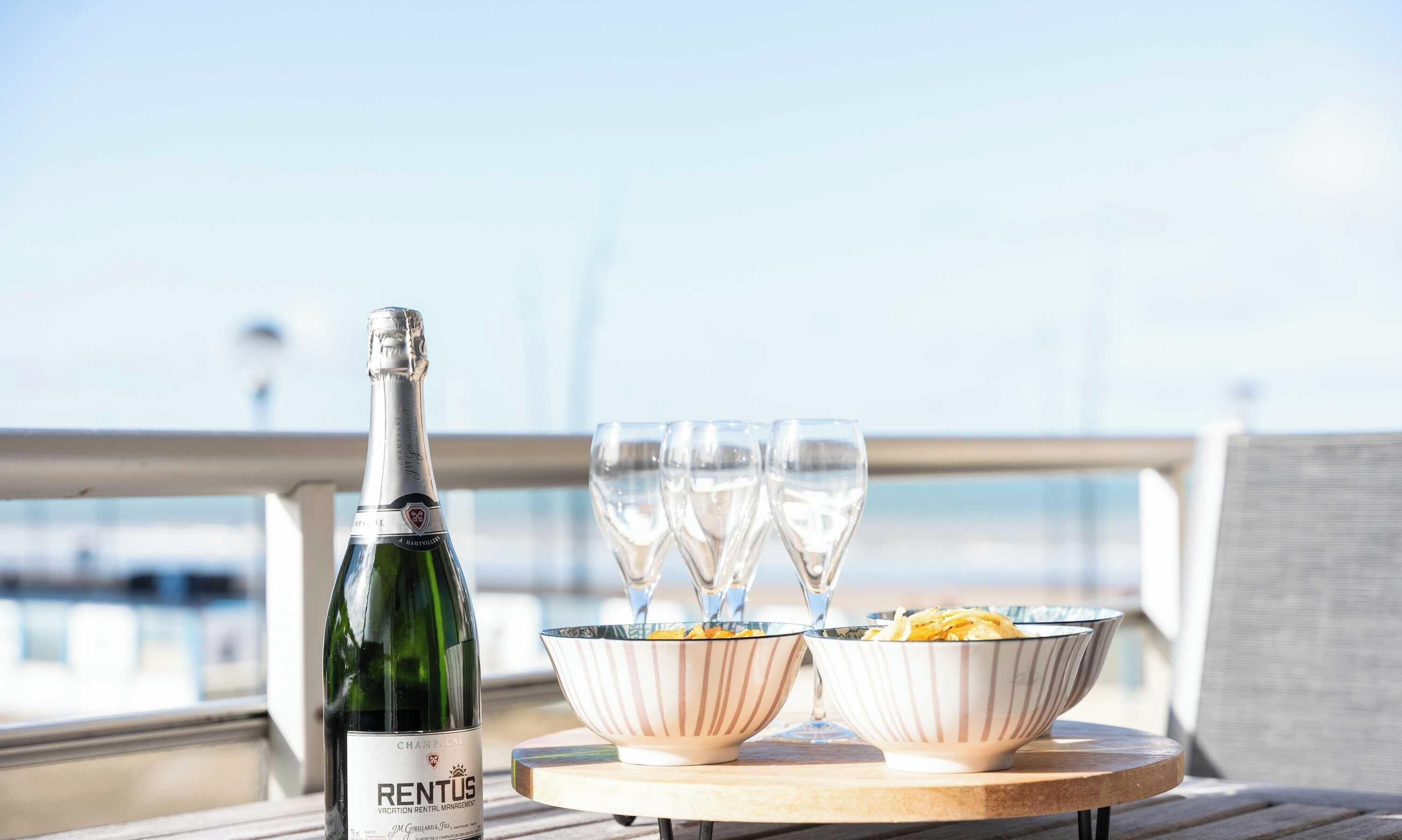 Eine Champagnerflasche, drei Gläser und zwei Schalen auf einem Tisch mit Meerblick.