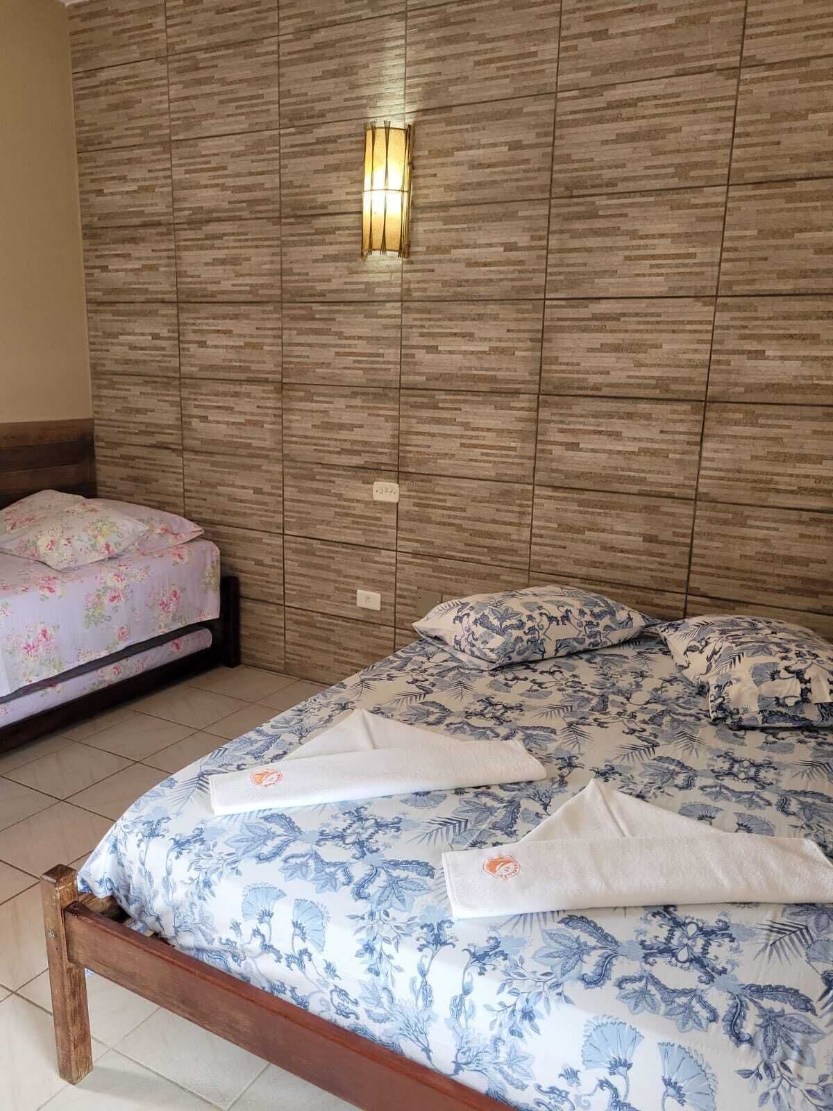 Quarto com cabeceira de madeira, cama com roupa de cama floral e mesa de cabeceira com colcha floral rosa.