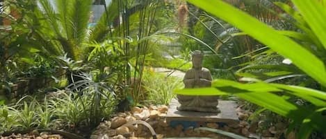 Un jardín con una estatua de Buda, rodeado de plantas tropicales y guijarros.