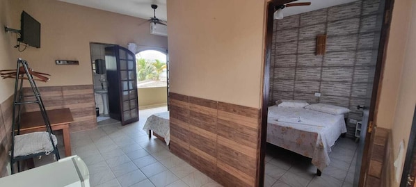 A bedroom with a wooden headboard, a bed with a floral bedspread, a small table, and a chair. - PousadaBoramar (Bertioga)