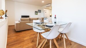 Un comedor moderno con una mesa de vidrio y sillas blancas, un sofá al fondo y una botella de vino sobre la mesa.