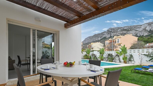 A patio with a round table and chairs, a glass door leading to an indoor area, and a pool with a view of buildings and mountains. - Limoncello - Denia, Costa Blanca (Denia)