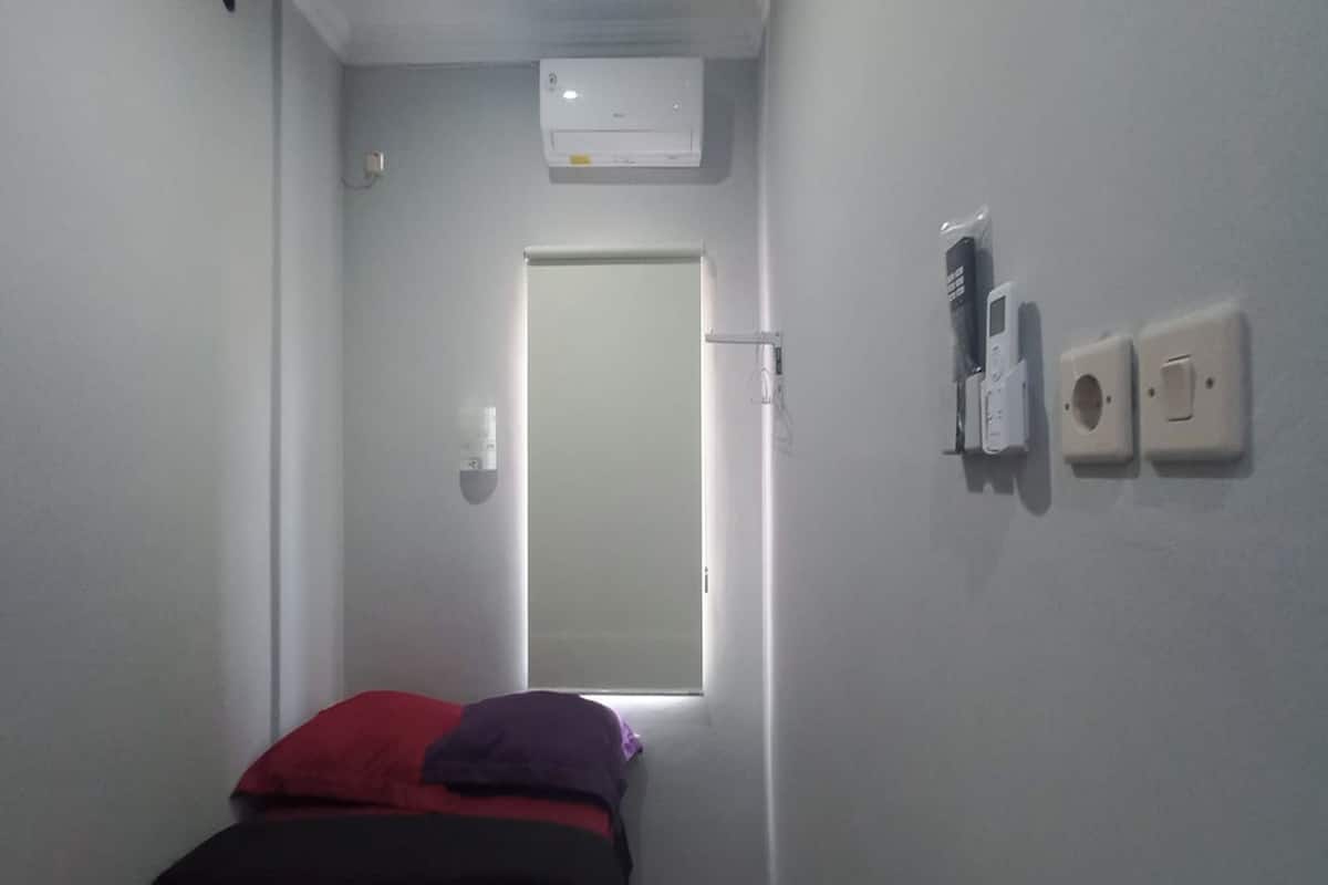 A narrow hallway leading to a door, with a bed at the end. There is an air conditioning unit mounted on the wall above the door.