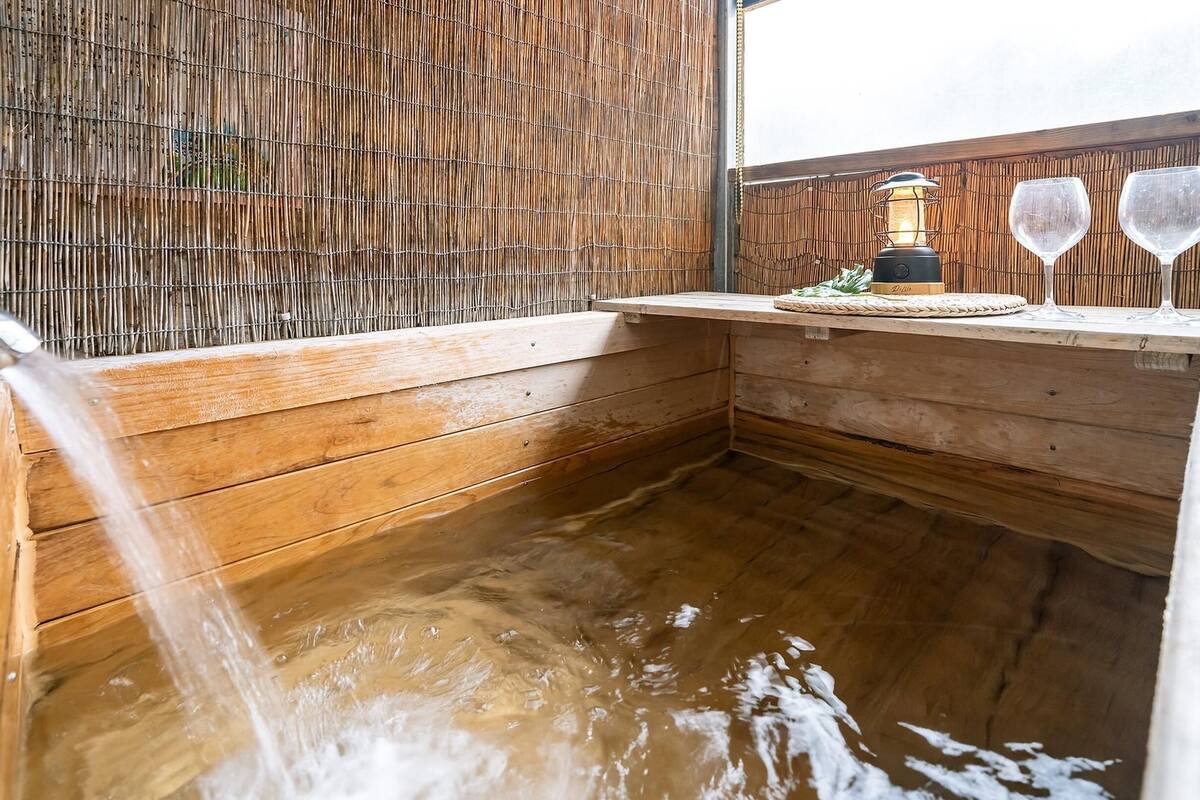 Ein hölzernes Whirlpool mit Bambuswand, einem Wasserhahn und zwei Weingläsern auf einer Holzablage.