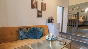 Un salon moderne avec un canapé marron, des coussins à motifs, une table basse en verre et un coin cuisine équipé d’un réfrigérateur et d’une machine  à café.