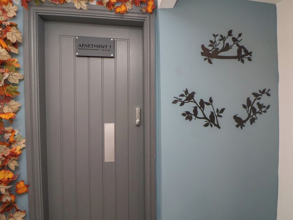 Una puerta con el letrero "APARTMENT 1" y una cerradura electrónica, rodeada de hojas otoñales.