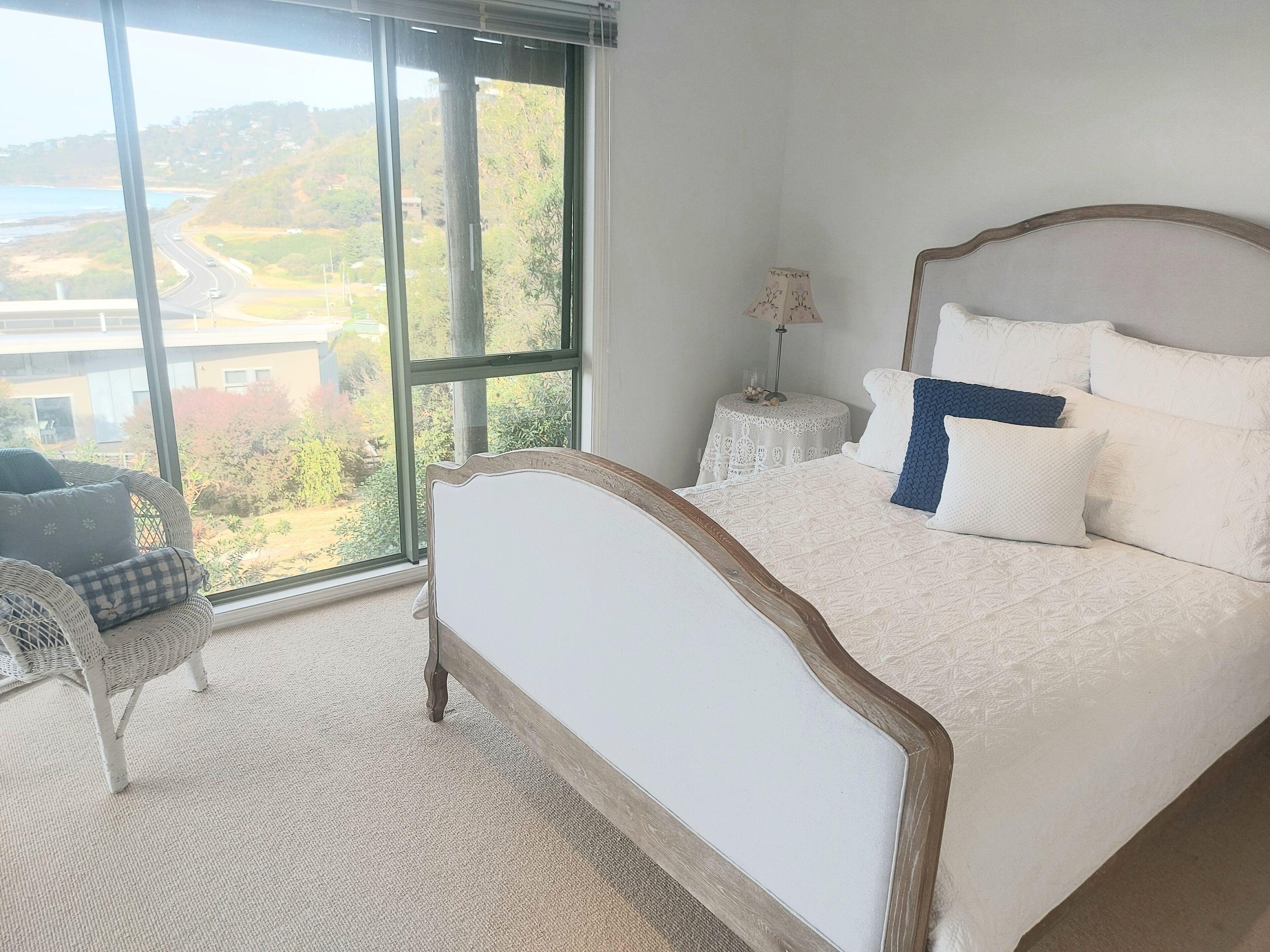 A bedroom with a large window, a bed with white bedding, a chair, and a small table with a lamp.