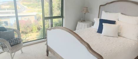 A bedroom with a large window, a bed with white bedding, a chair, and a small table with a lamp.