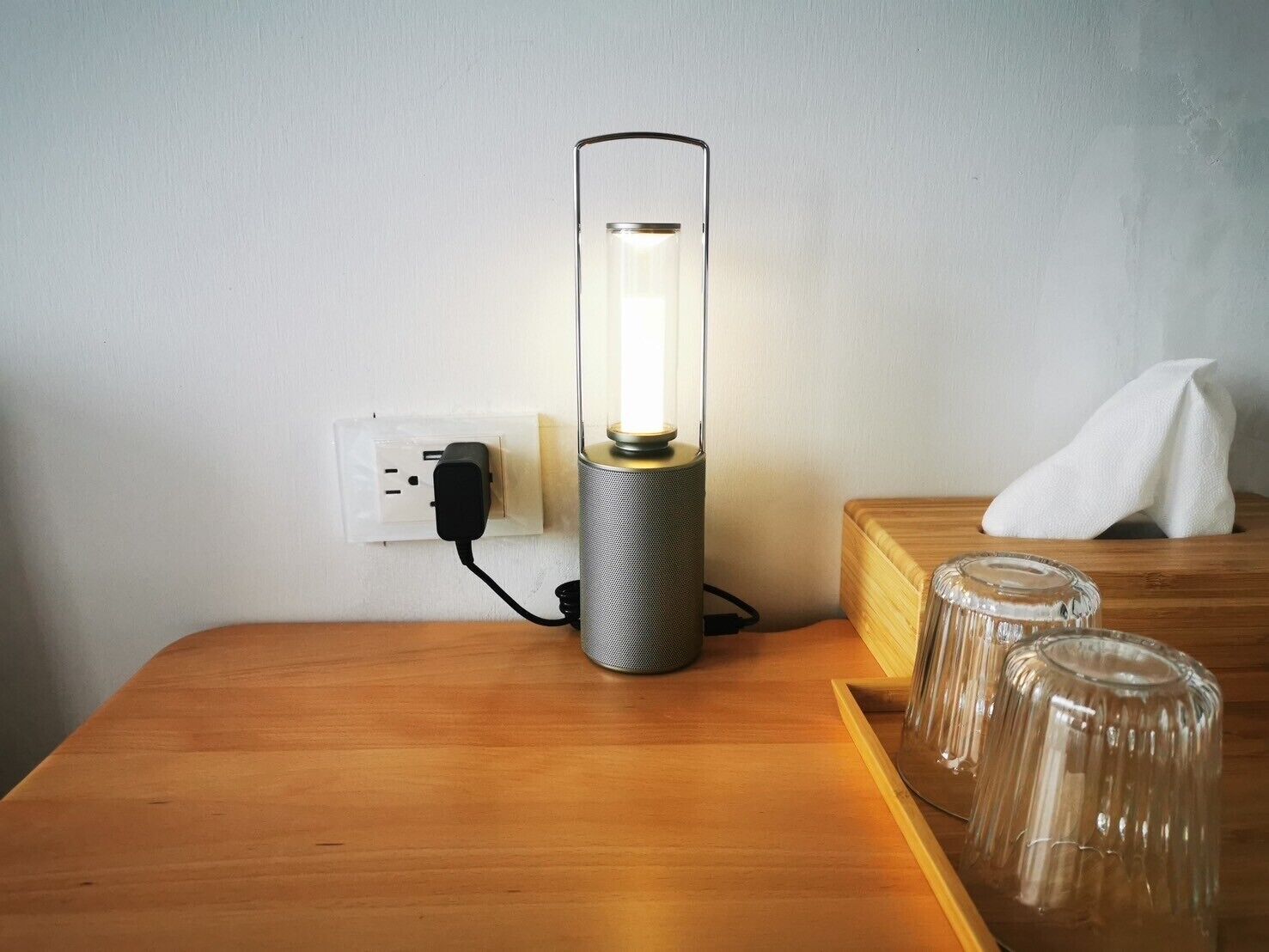 Una mesita de noche con una lámpara, un dispensador de pañuelos y un recipiente de vidrio.