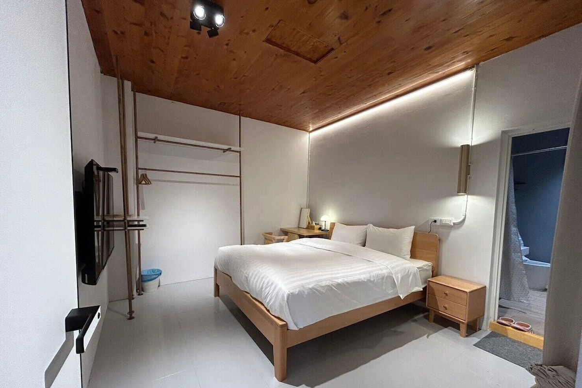 A modern bedroom with a wooden ceiling, a bed with white bedding, bedside tables, a wall-mounted TV, and a window with a view.