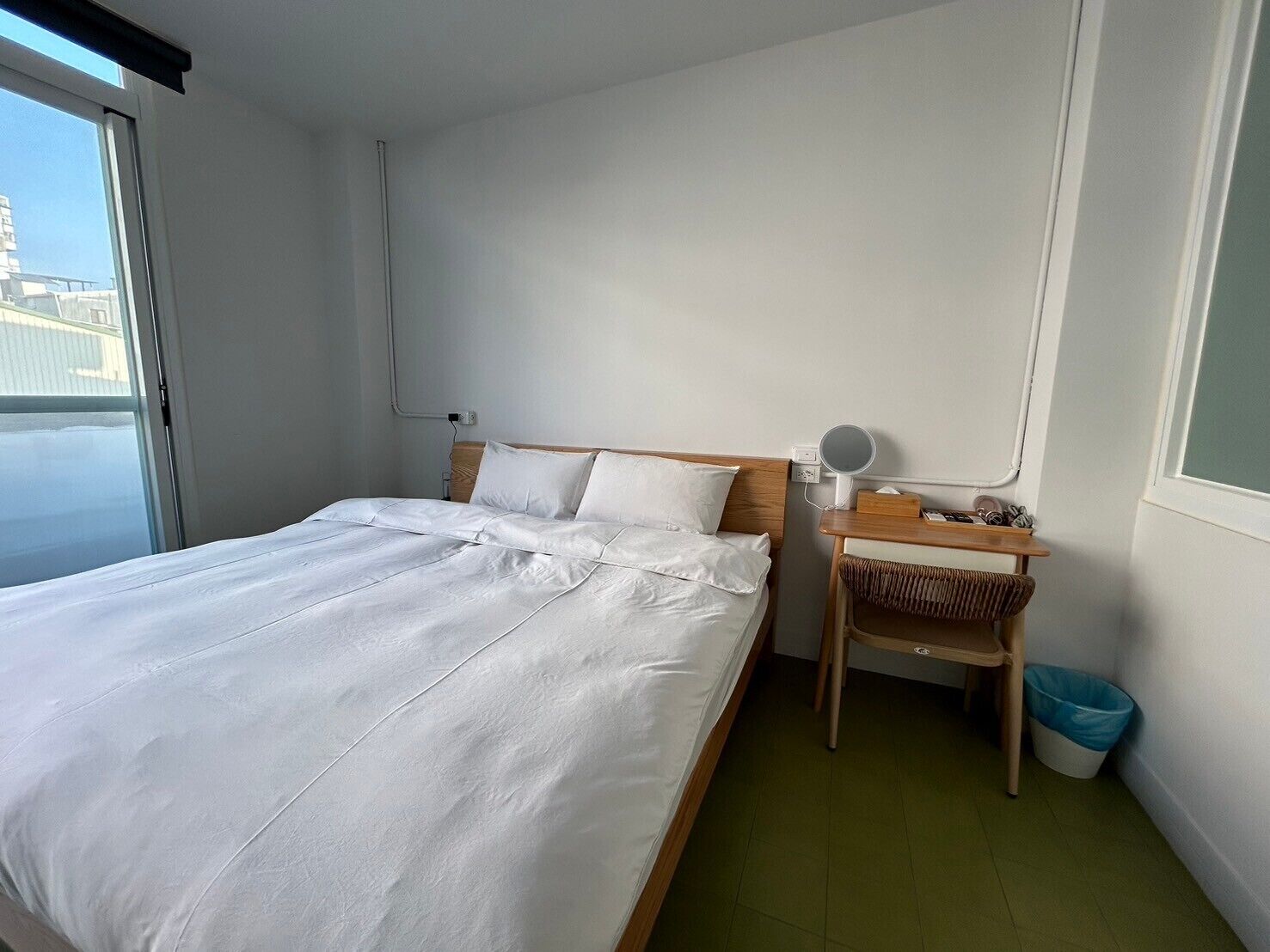 A neatly made bed with white linens, a small bedside table with a mirror, and a trash bin.