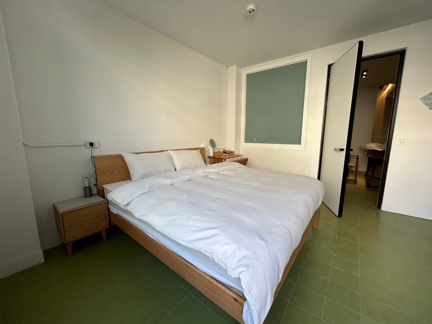 Ein ordentlich bezogenes Bett in einem Zimmer mit grünem Boden, einem Nachttisch mit Lampe und einem großen Fenster.