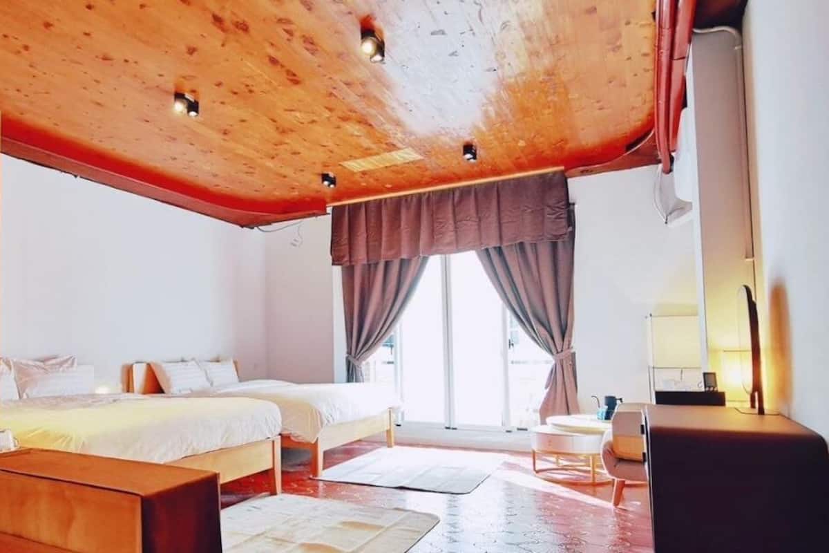 A bedroom with a wooden ceiling, two beds, a bedside table, a chair, and a window with curtains.
