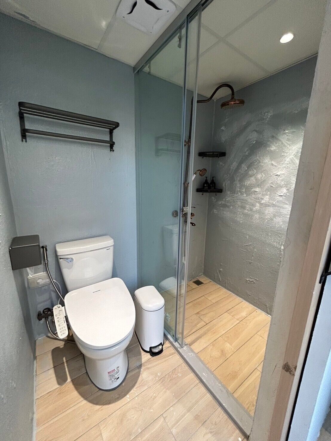 Un baño con inodoro, un cubo de basura, cabina de ducha de vidrio y repisa sobre el inodoro.