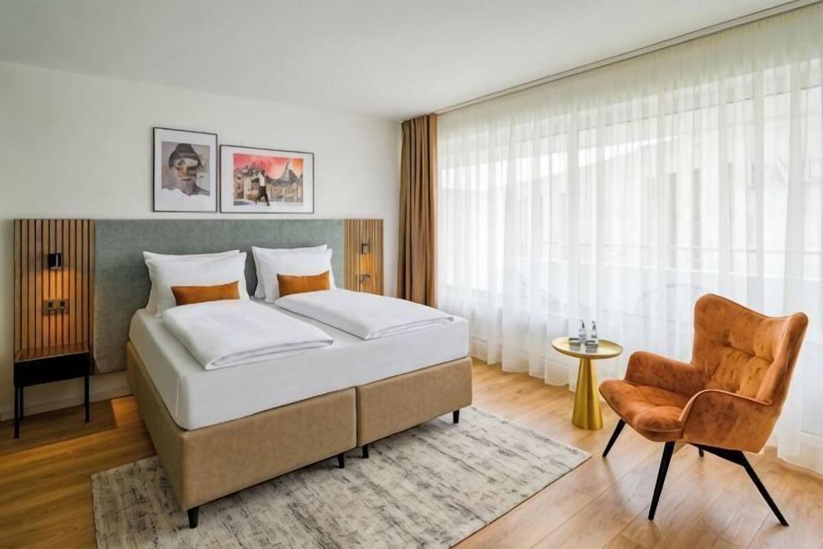 Une chambre d’hôtel moderne, dotée d’un grand lit, de deux tables de chevet, d’une chaise et offrant une vue sur la ville à travers de fines rideaux.