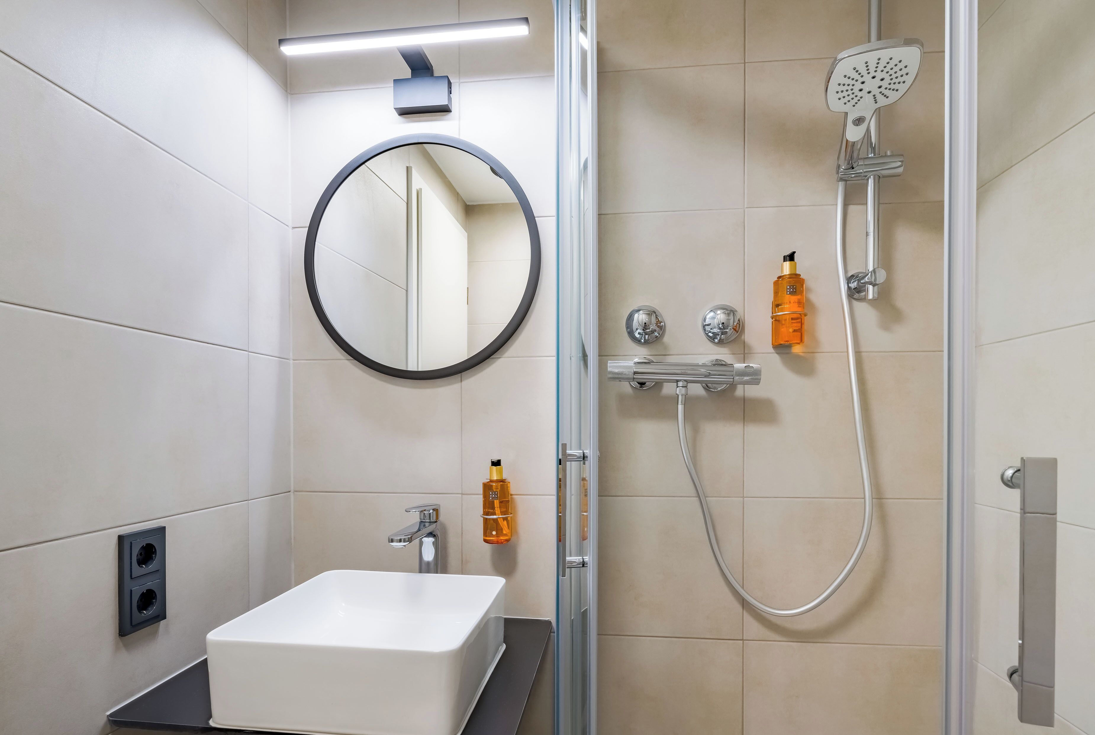 A modern bathroom with a white sink, a round mirror, a handheld showerhead, and two bottles of shampoo.