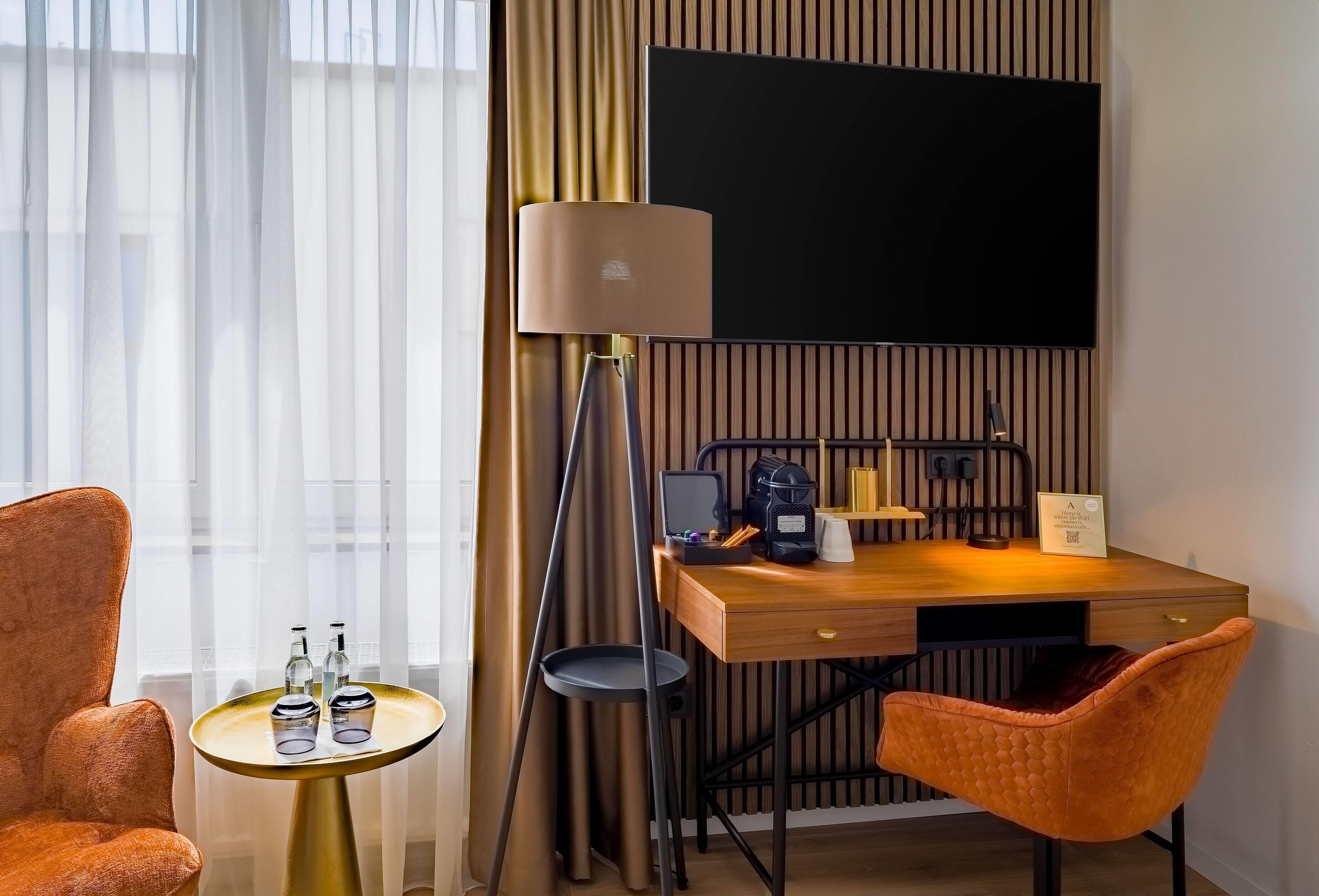 A hotel room with a wooden desk, a lamp, a TV, and two chairs.