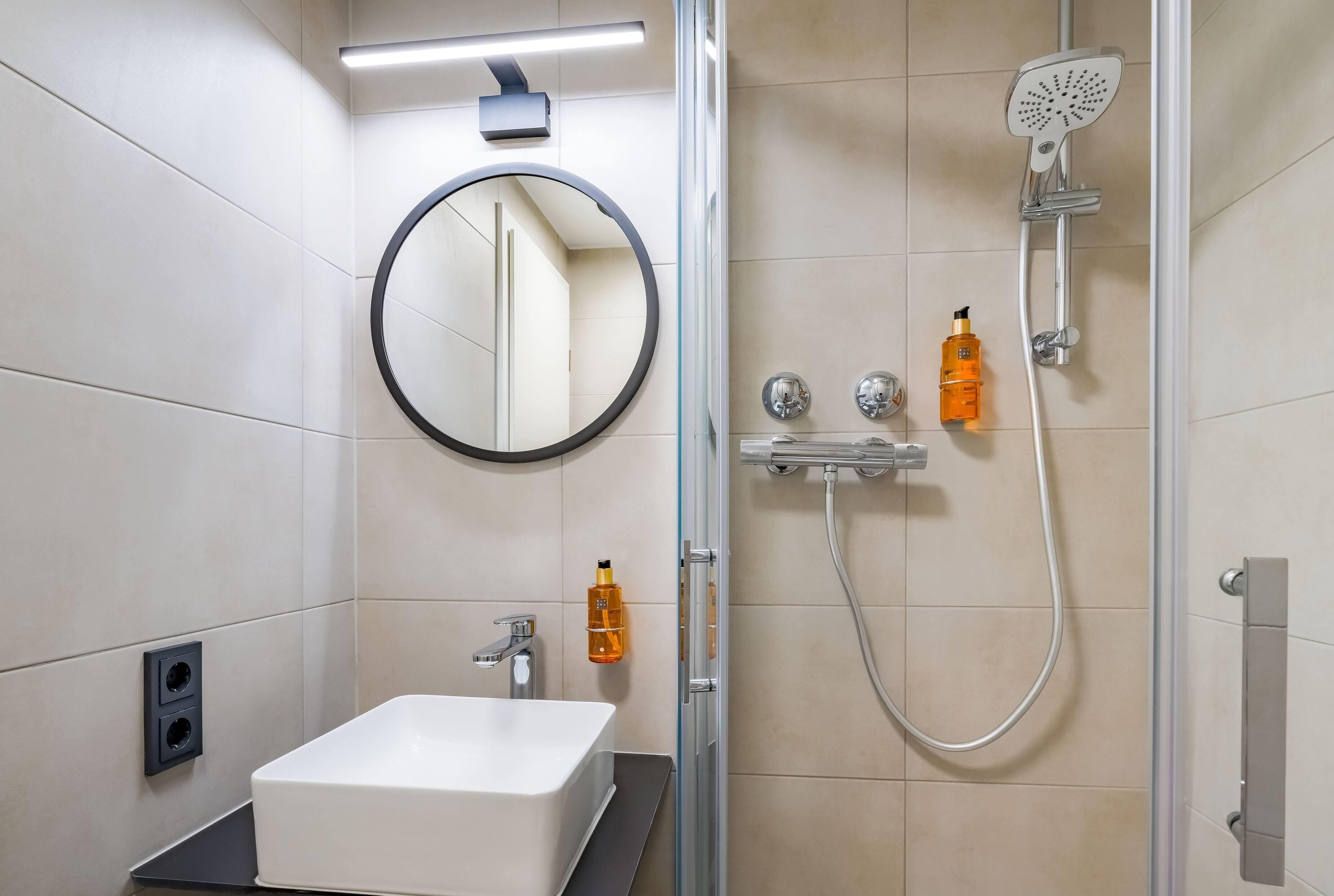 Een moderne badkamer met een wit wastafel, een ronde spiegel, een handdouche en twee zeepflessen.