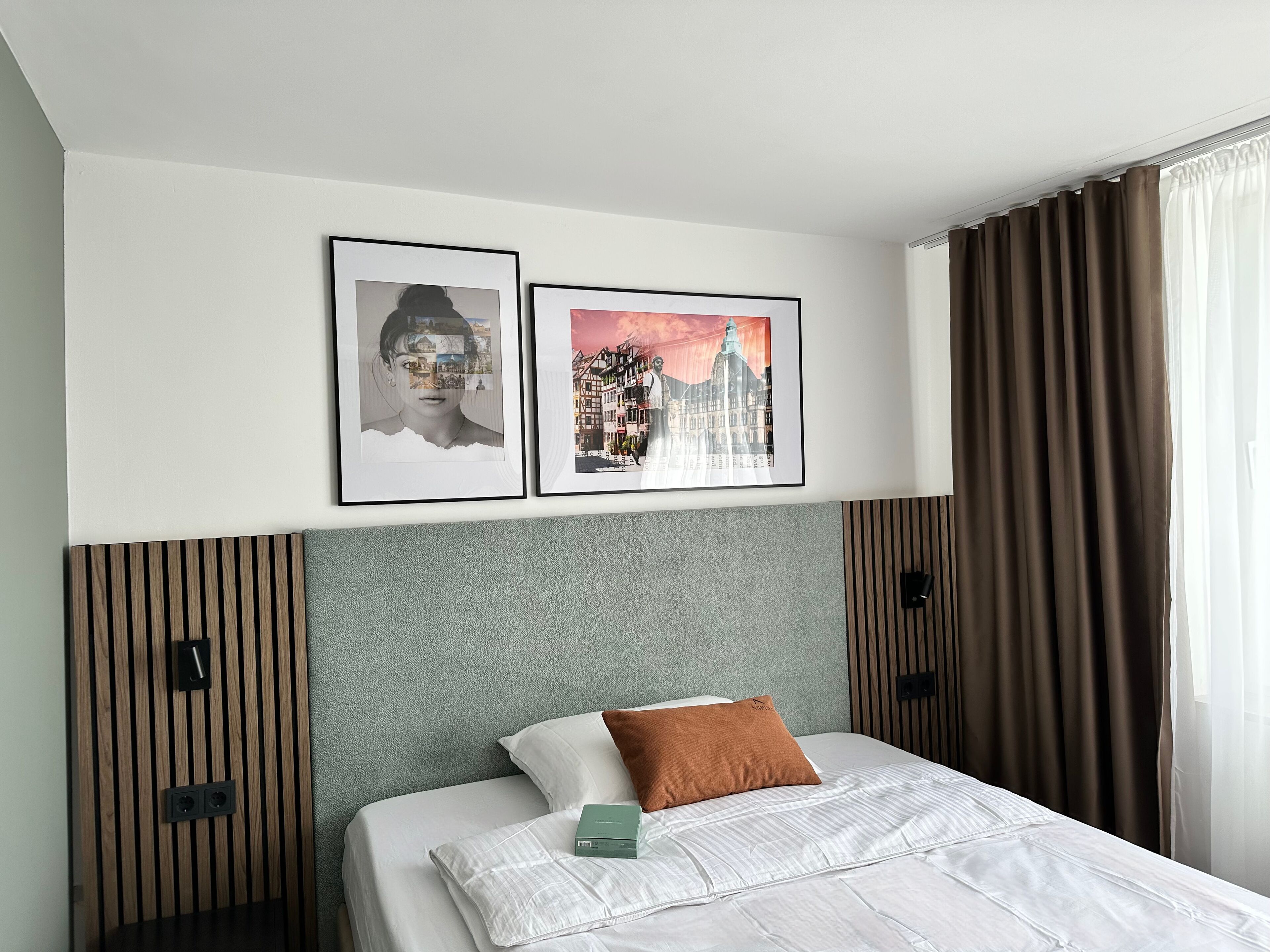A neatly made bed with a white comforter and a brown pillow, a framed picture of a cityscape, and a framed portrait on the wall.